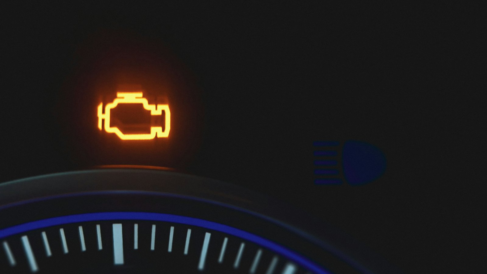Amber check engine warning light glowing on a dark dashboard next to a blue high-beam indicator