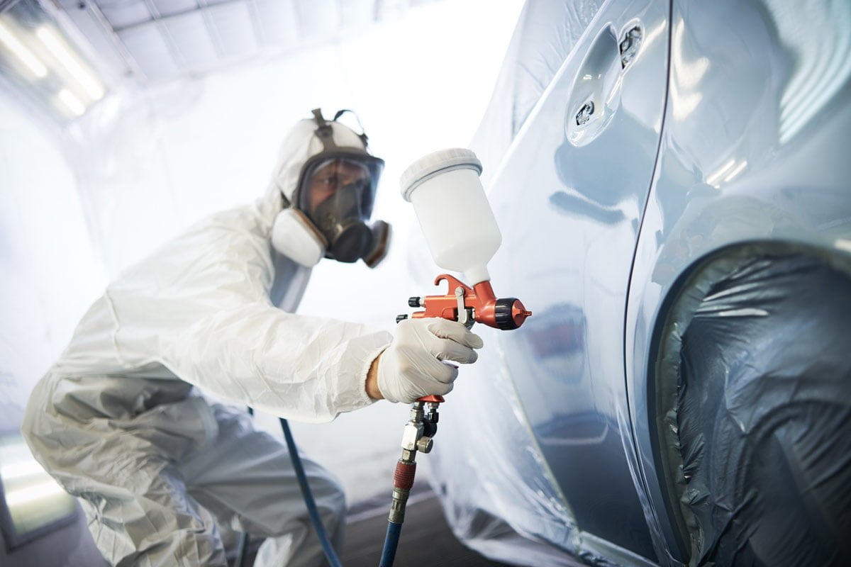 Body technician in protective gear refinishing a car panel with a spray gun
