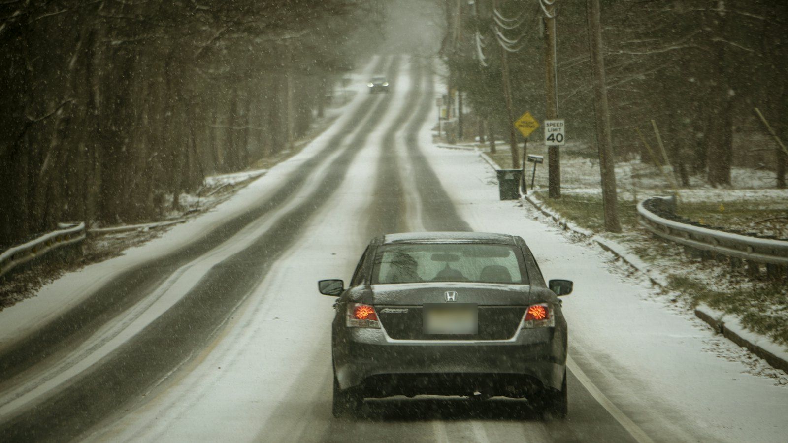 Sedan with its brake lights on, slowing down on a snow-covered two-lane road lined with trees, the kind of salted winter driving New Hampshire brakes deal with for months