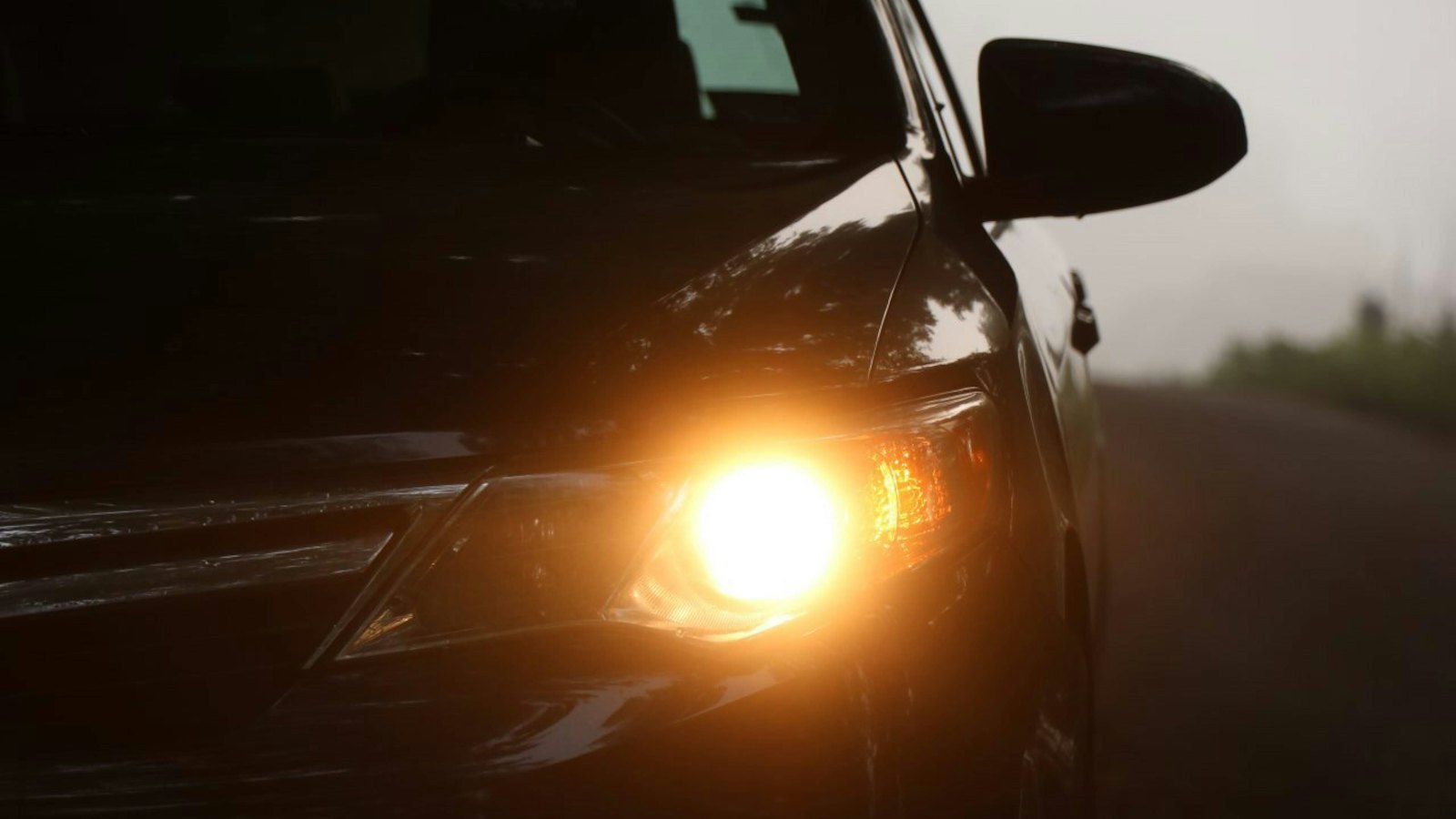 Dark sedan with its headlight on at dusk on a quiet road, a quick lighting check any driver can do without a state inspection