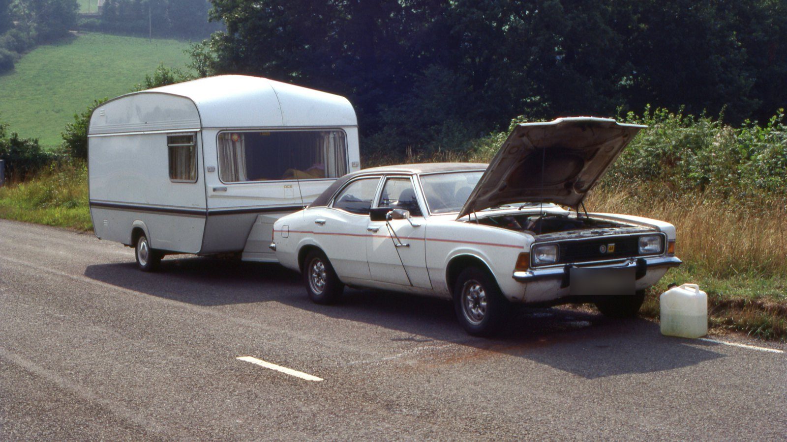 Sedan towing a small camper, pulled over on the roadside with its hood raised and a jug of water on the ground, the classic warm-weather overheating stop