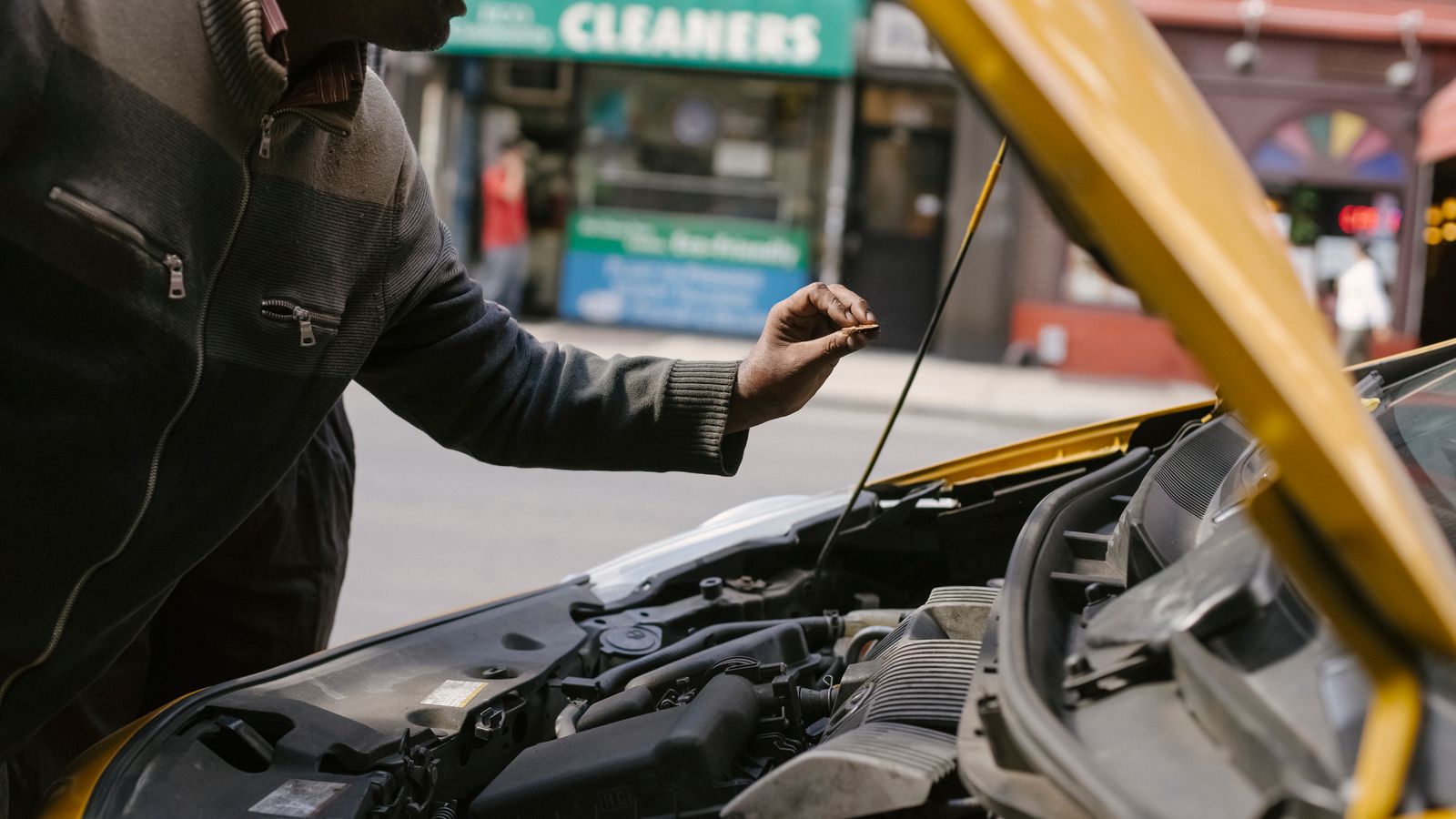 Man checking the engine oil dipstick under the open hood of a yellow car parked on a city street