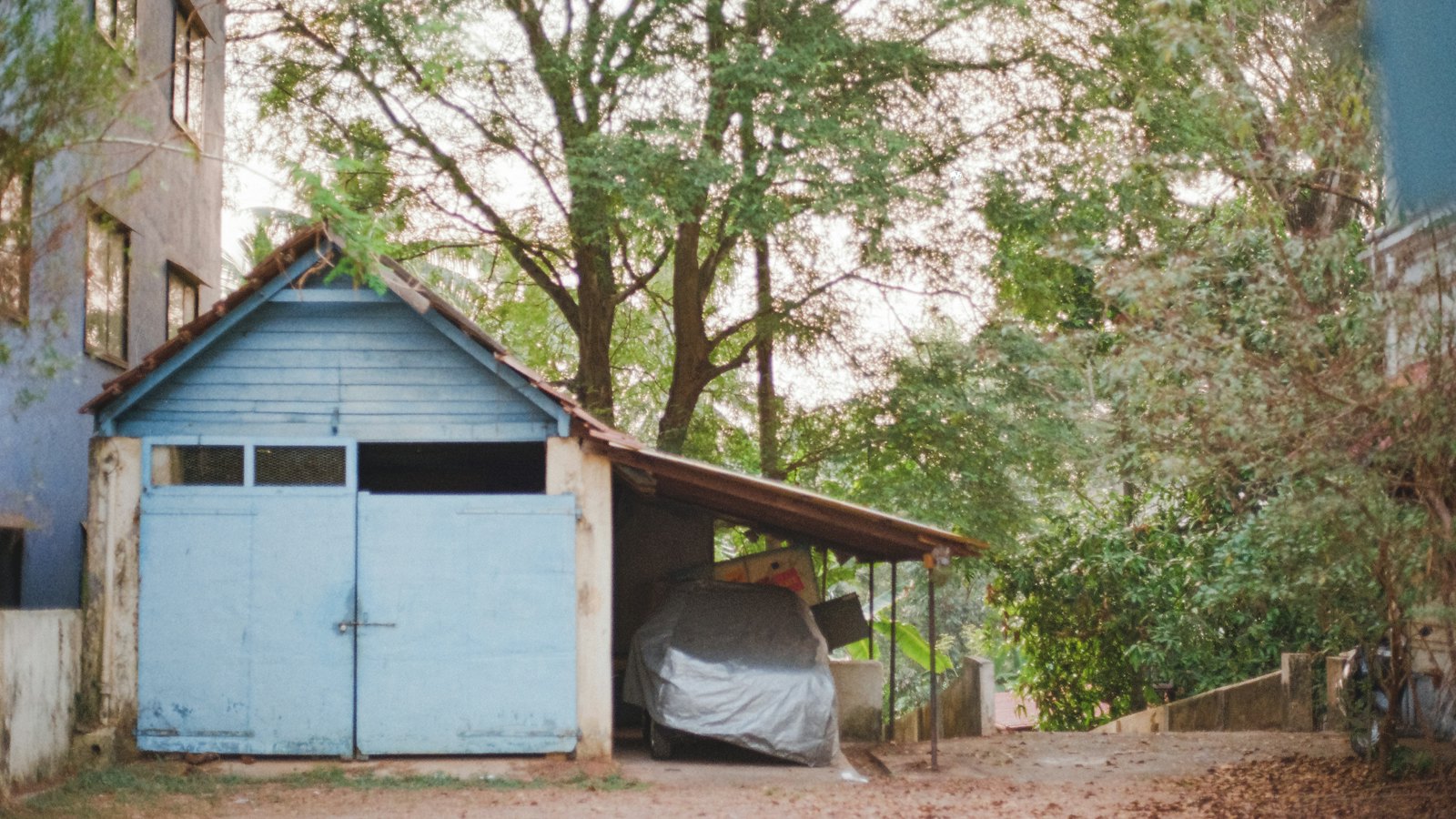 Car under a fitted cover parked beneath a carport next to an old garage, one answer to how to store a car for winter
