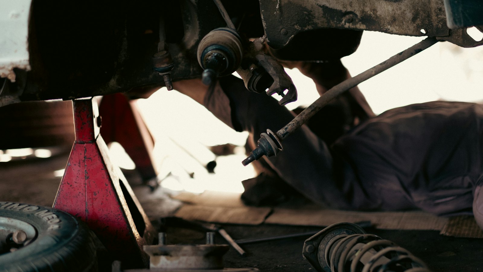 Mechanic lying under a car raised on red jack stands, tracing a fluid leak back to its source