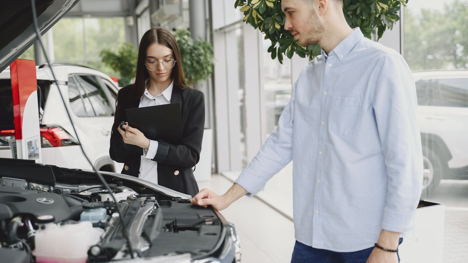 Car buyer and a salesperson looking under the open hood of a car in a dealership showroom, the moment to plan a post-purchase car inspection