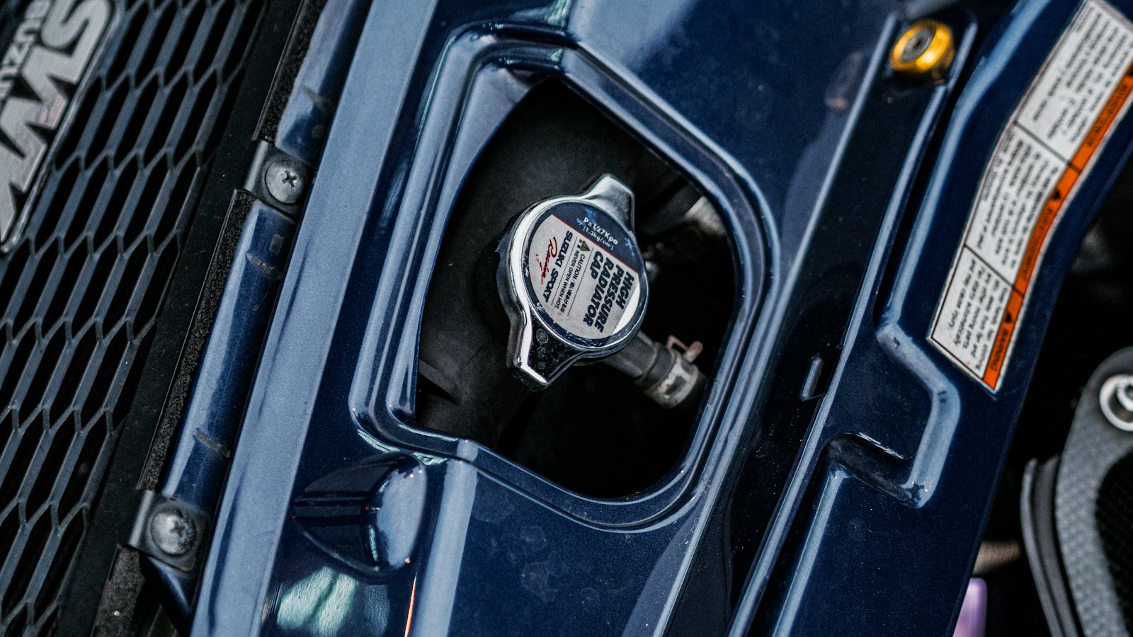 High-pressure radiator cap on a car's radiator neck beside the grille, which should never be opened while the engine is hot