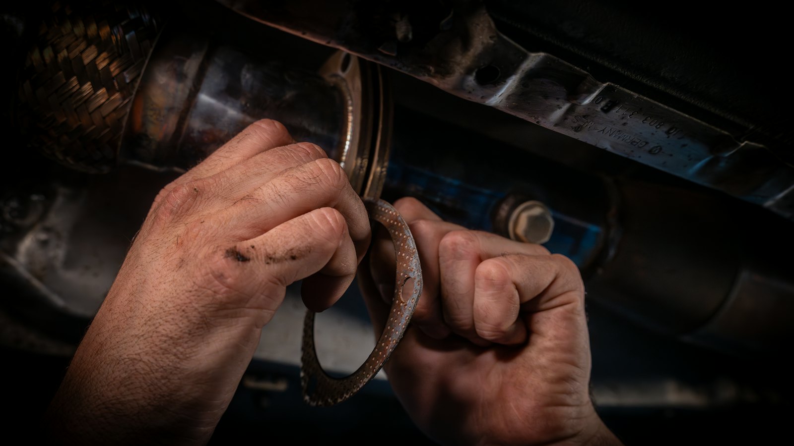 Technician's hands pulling an old gasket from a rusted exhaust flange under a car during catalytic converter removal