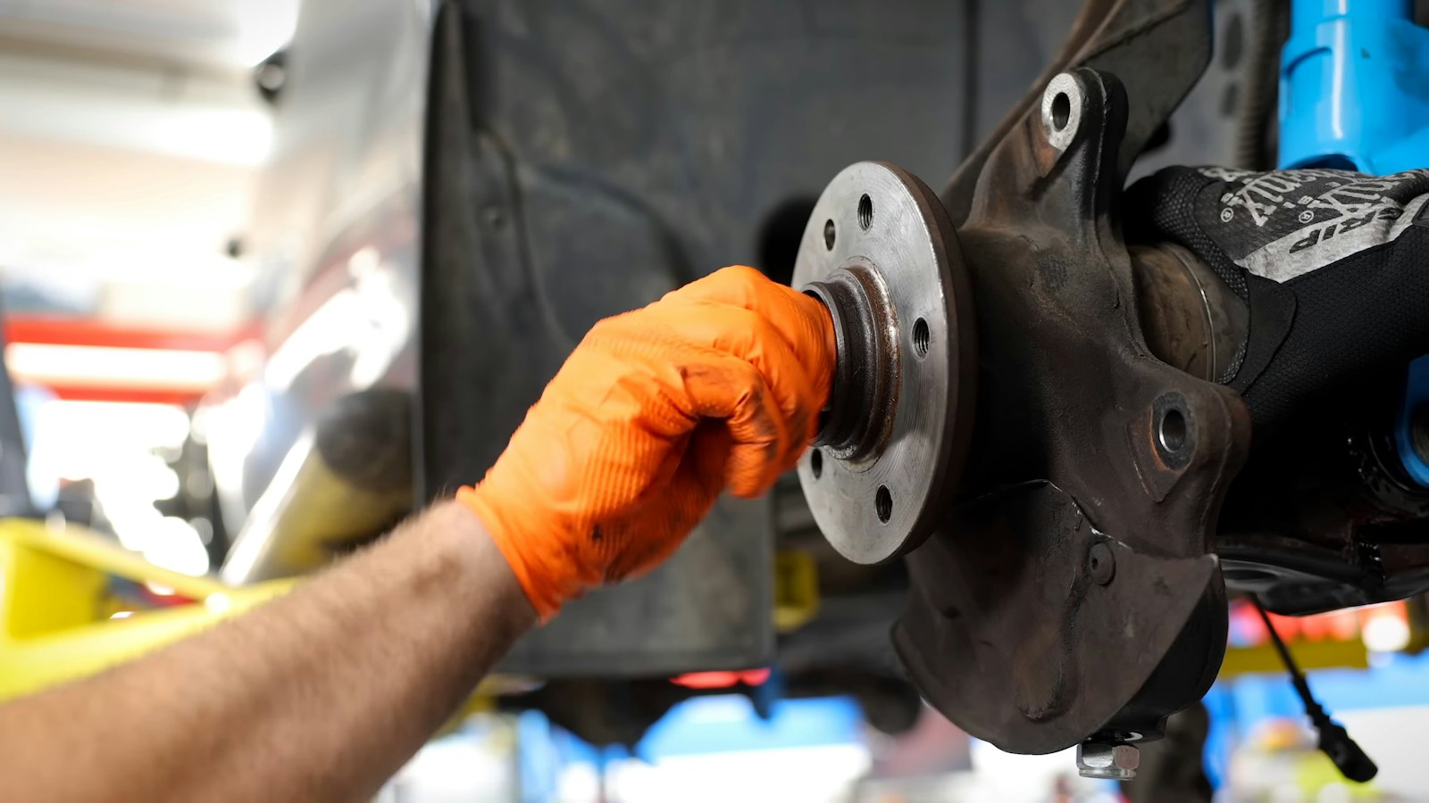 Technician in an orange glove steadying a corroded wheel hub and steering knuckle on a car raised on a shop lift, with the brake rotor removed